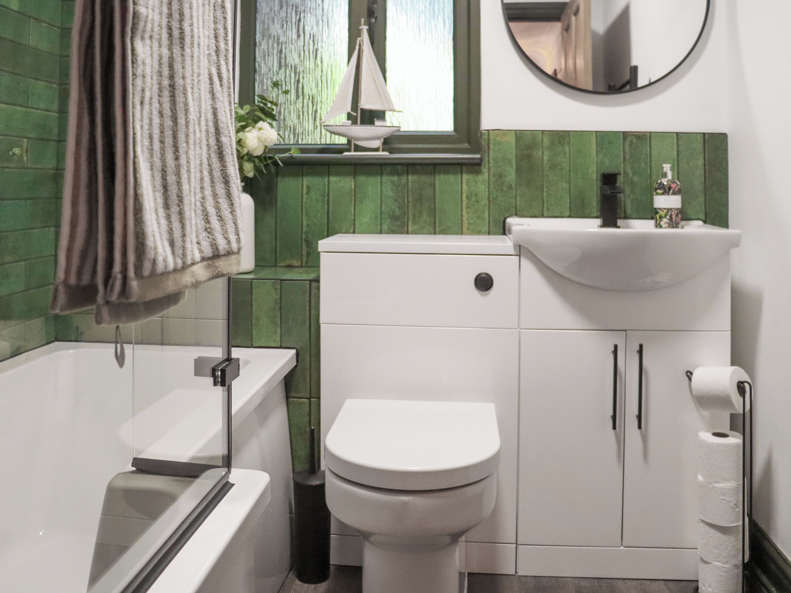 A bathroom with a white toilet, a white sink, and a bathtub. There is a green tiled wall and a small sailboat decoration on the window sill.