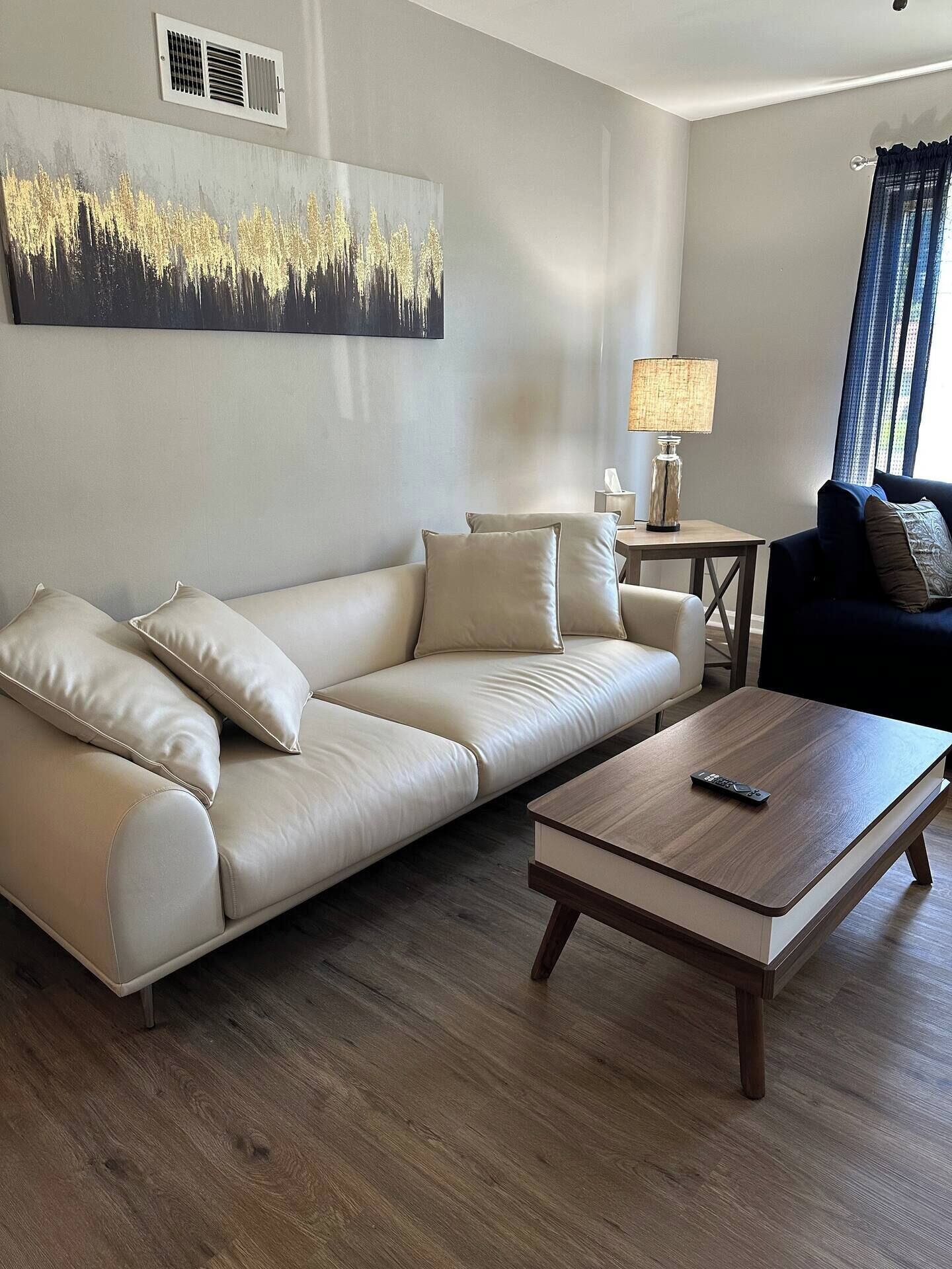 Un salon moderne avec un canapé en cuir beige, une table basse en bois et un fauteuil bleu foncé. Un tableau est accroché au mur, et une lampe est posée sur une petite table.