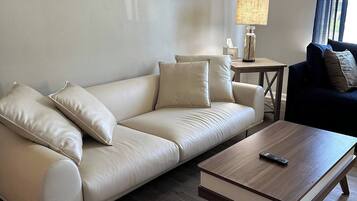 Un salon moderne avec un canapé en cuir beige, une table basse en bois et un fauteuil bleu foncé. Un tableau est accroché au mur, et une lampe est posée sur une petite table.