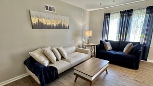 Ein Wohnzimmer mit einem beigefarbenen Sofa, einem schwarzen Sofa, einem Couchtisch aus Holz und einer Lampe auf einem Beistelltisch.