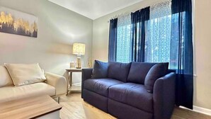 Ein Wohnzimmer mit einem dunklen Sofa, einem hellen Sessel, einem kleinen Beistelltisch mit einer Lampe und einem Gemälde an der Wand.