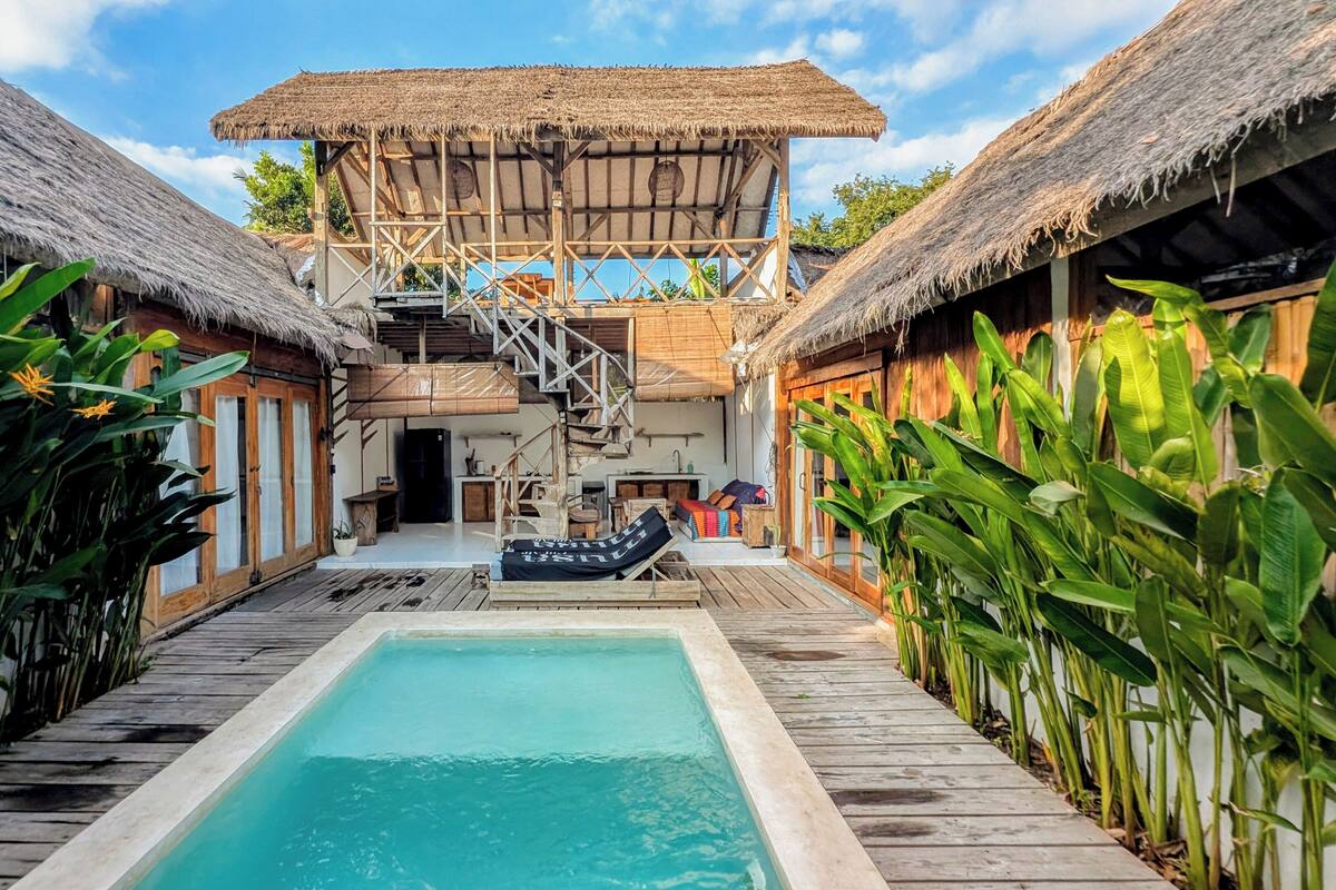 Un espace piscine doté d’une terrasse en bois, d’une structure à toit de chaume et de plantes tropicales.