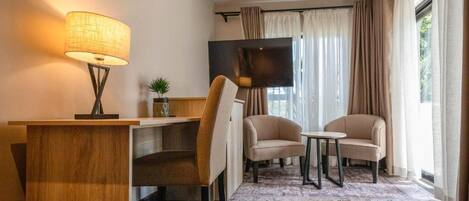 A modern hotel room with a wooden desk, two armchairs, a small table, a TV, and a large window with curtains.