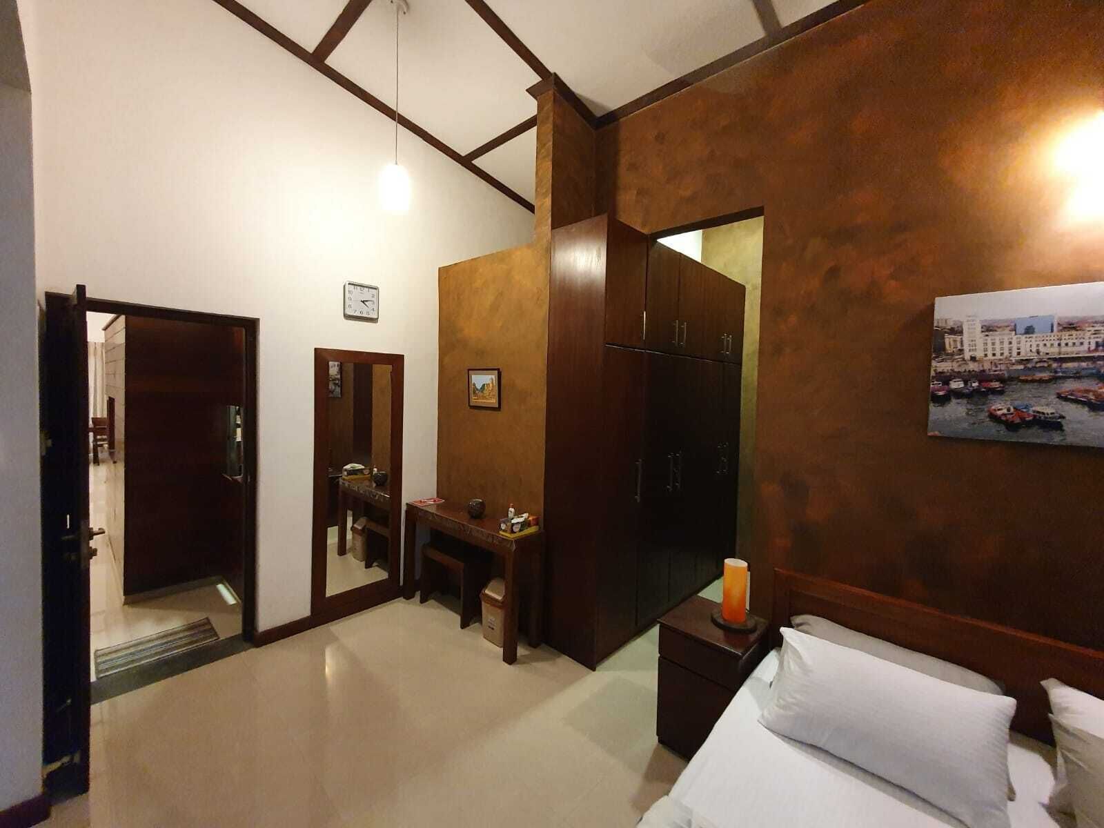 A hotel room with a bed, a nightstand, a mirror, a wooden wardrobe, and a painting of a harbor scene.