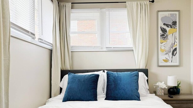 A neatly made bed with a teal blanket and two blue pillows, a white towel, and a framed floral artwork on the wall.