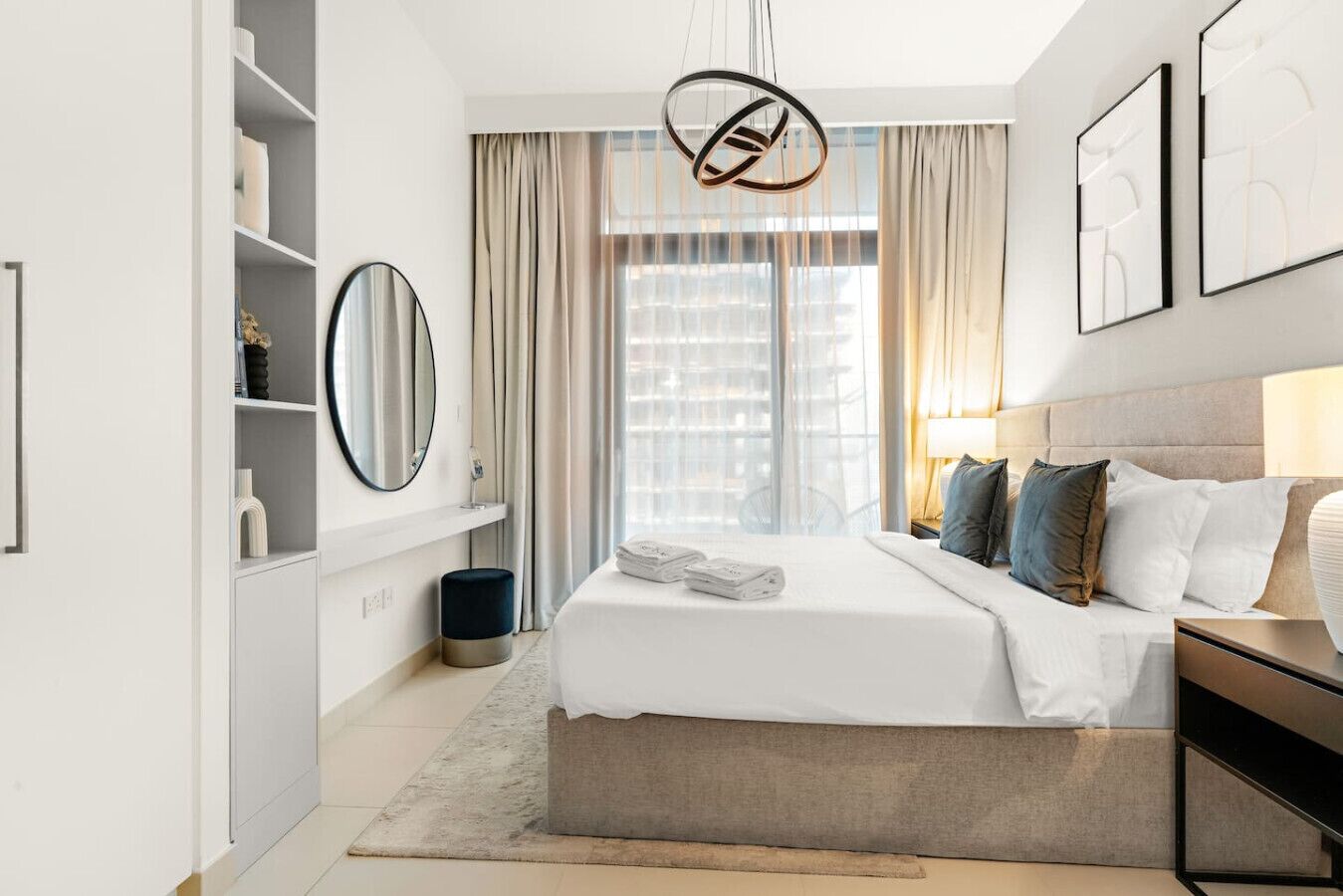 A modern hotel room with a large bed, a bedside table, a mirror, and a view of the city through large windows.