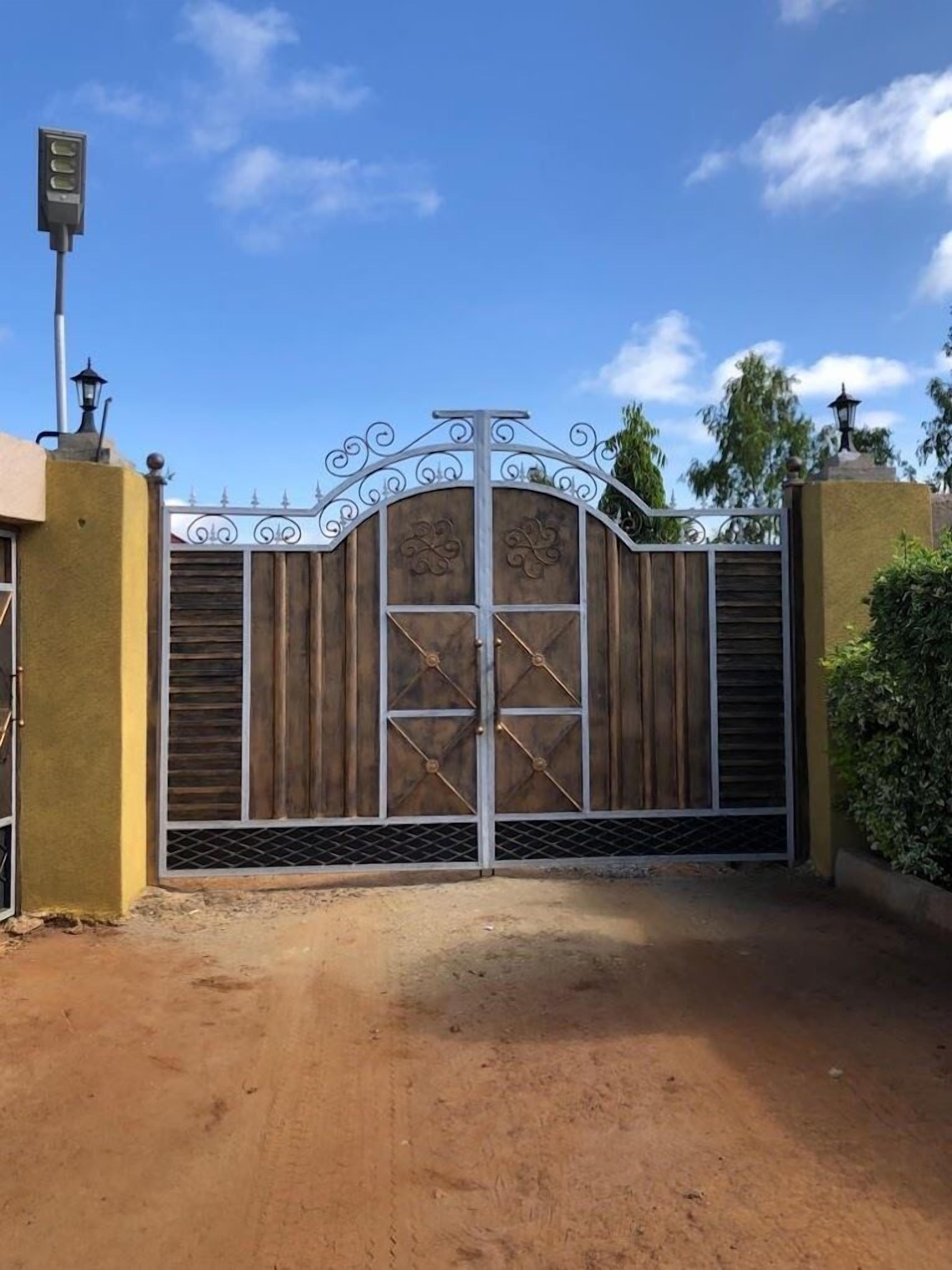Una entrada protegida por un portón metálico con detalles decorativos, que conduce a un edificio con un muro amarillo y un farol.
