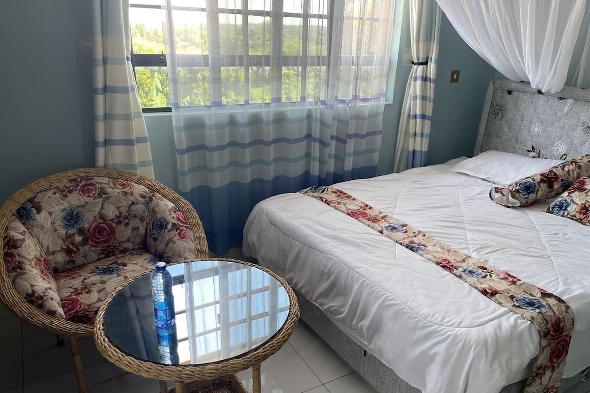 Un dormitorio con cama, una silla de mimbre, una mesita redonda y una ventana con cortinas.