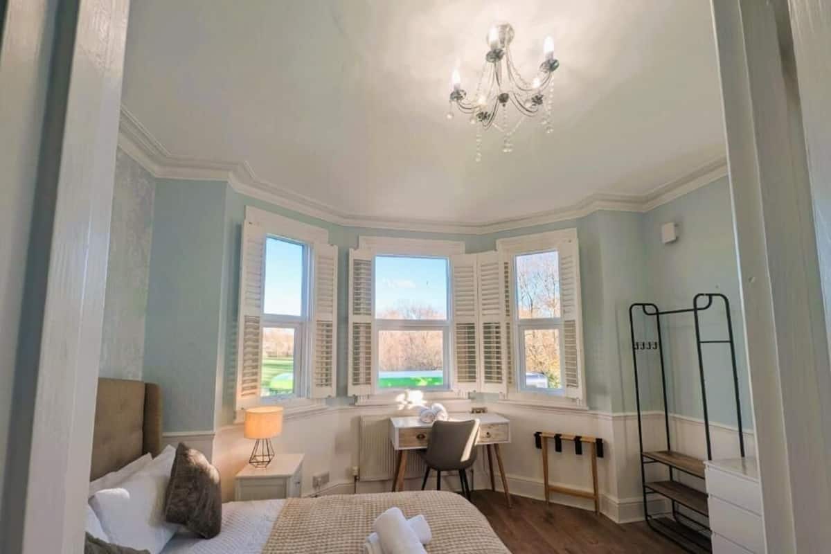 A bedroom with a bed, a desk, and a chair. There are windows with shutters, a radiator, and a chandelier.