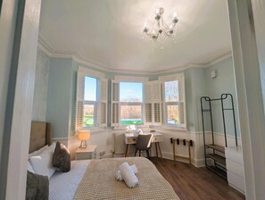 Een slaapkamer met een bed, een bureau en een stoel. Er zijn ramen met luiken, een radiator en een kroonluchter.