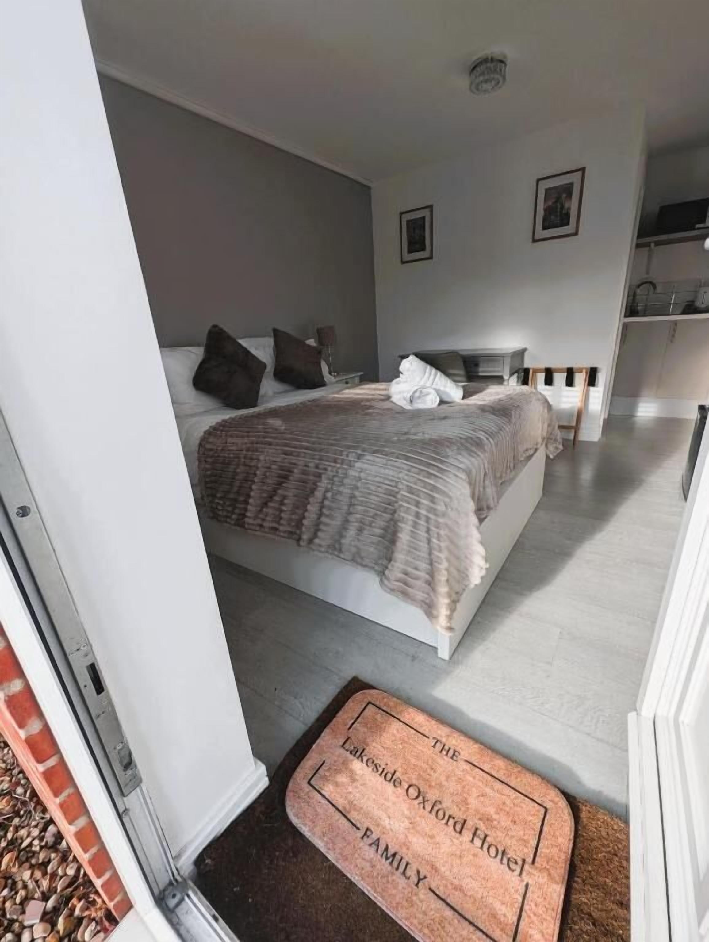 Une chambre à coucher avec un lit, des oreillers, une table de chevet et un petit bureau. Il y a une porte avec un paillasson sur lequel on peut lire : « Lakeside Oxford Hotel, FAMILLE ».
