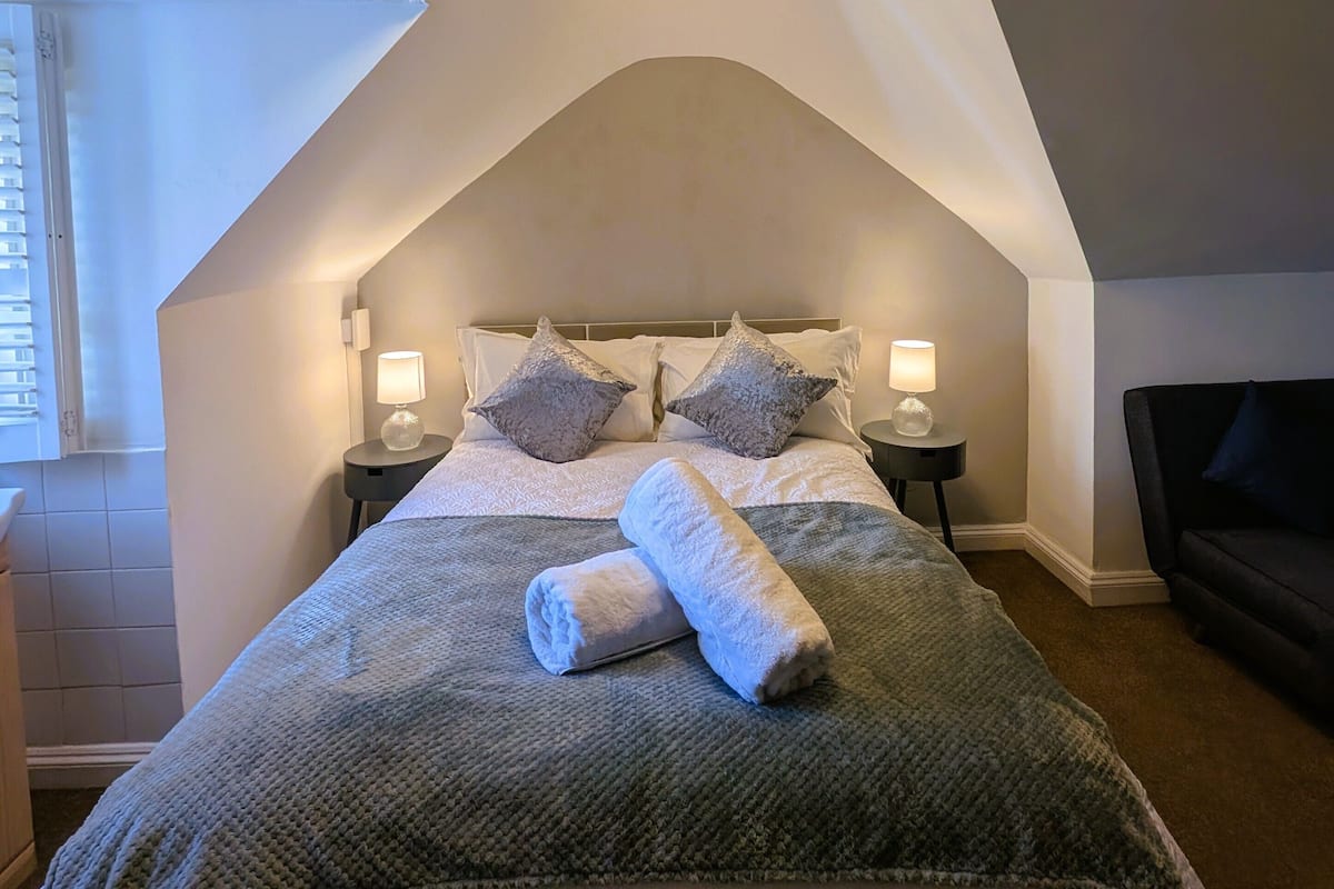 A bedroom with a high ceiling, a large bed with a rolled towel, two bedside lamps, and a wall-mounted light fixture.