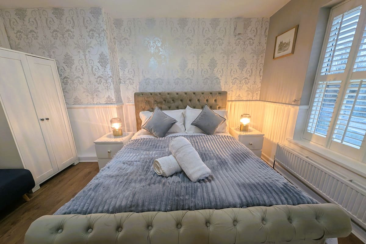 A bedroom with a tufted headboard, a large bed with a grey quilt, two bedside lamps, a white wardrobe, and a window with blinds.