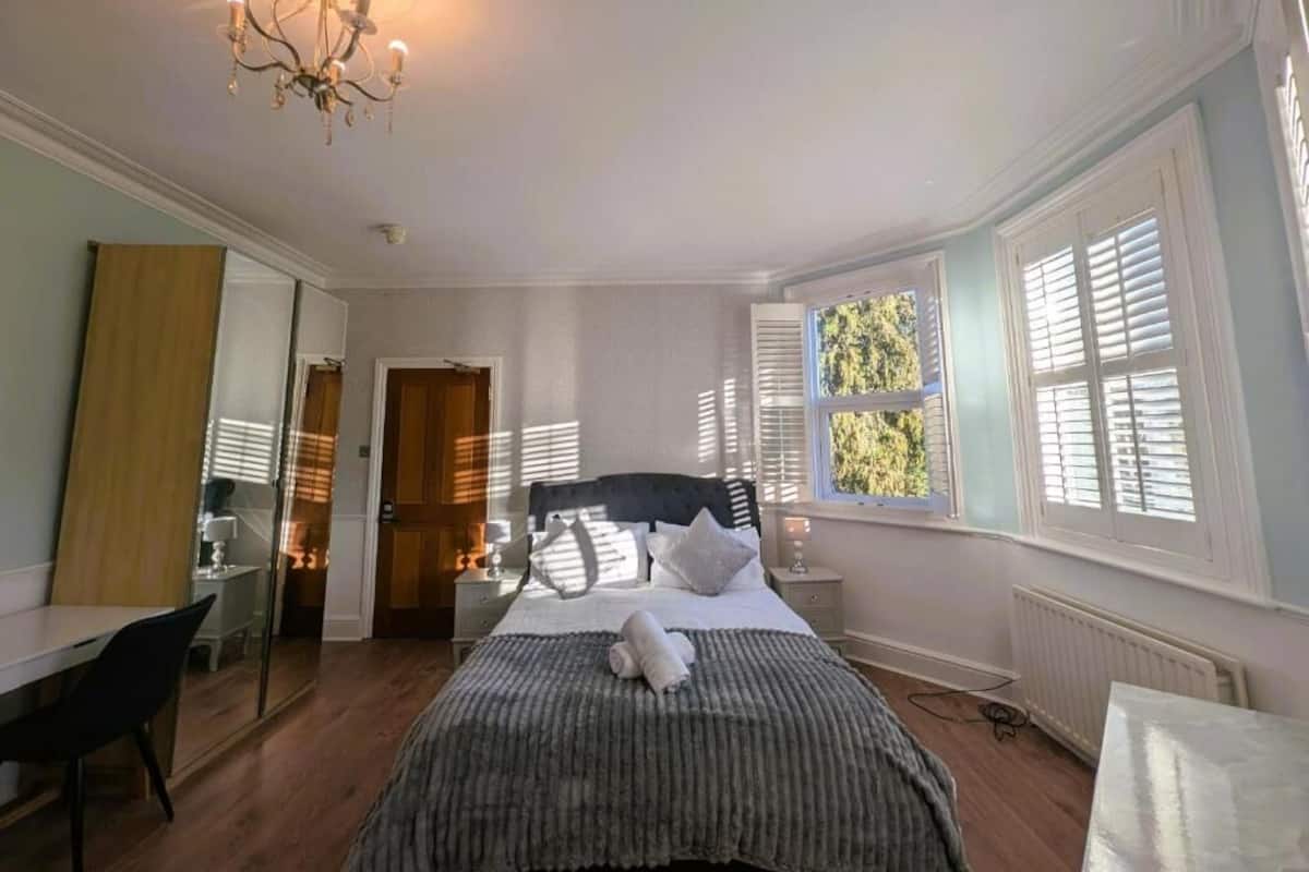 A bedroom with a bed, bedside tables, a mirror, a chandelier, and a window with shutters.