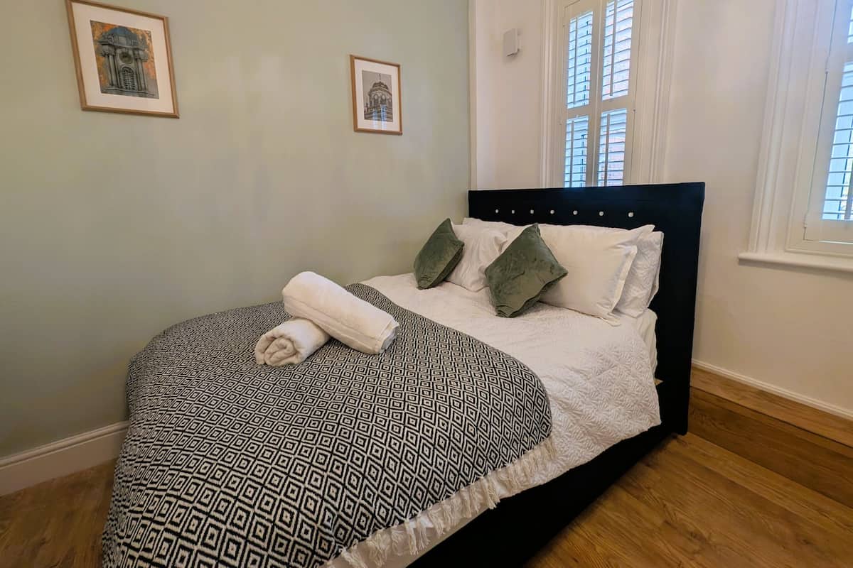 A neatly made bed with patterned bedding, two framed pictures on the wall, and a window with blinds.