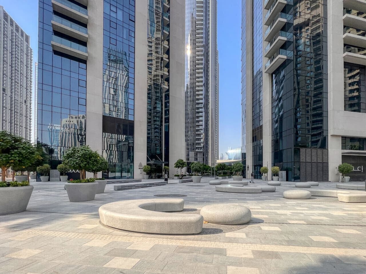A modern urban plaza with high-rise buildings, circular seating areas, and potted plants.