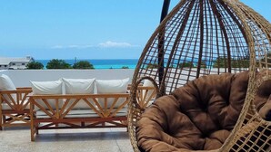 Une chaise suspendue avec un coussin marron, placée sur une terrasse carrelée, d’où l’on peut admirer un ciel bleu dégagé et l’océan.