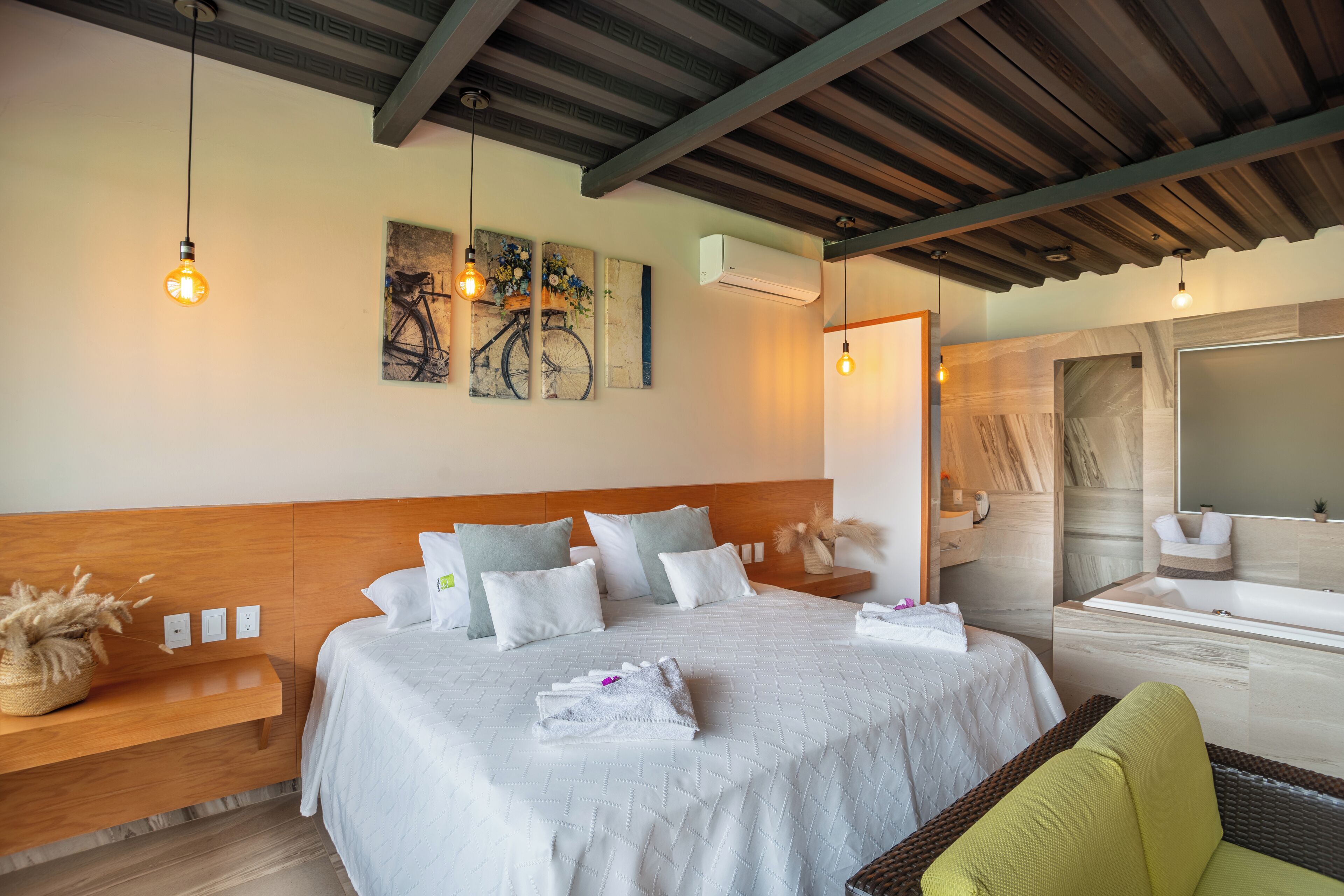 A hotel room with a large bed, a wooden headboard, a bench, a wicker chair, a bathroom with a bathtub, and artwork on the wall.