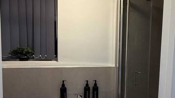 A bathroom with a white pedestal sink, a toilet, a shower with a glass door, and a window with vertical blinds.