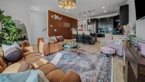 Un salon moderne avec un canapé, une table basse et un tapis. On aperçoit une cuisine à l’arrière-plan, avec des placards et des appareils électroménagers.