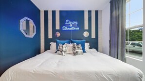 Une chambre à coucher avec un lit, un panneau lumineux néon sur lequel on peut lire « Denim & Rhinestones », et une fenêtre avec des rideaux.