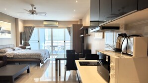 Ein modernes Hotelzimmer mit Bett, Schreibtisch, Stuhl, Deckenventilator, Klimaanlage, Küchenzeile mit Mikrowelle und Toaster sowie einem Balkon mit Tisch und Stühlen.