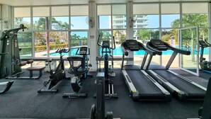 A gym with treadmills, stationary bikes, and weight machines. Large windows offer views of a swimming pool and palm trees. - Sea View Studio - Jomtien Beachfront - View Talay 8 (Muang Pattaya)