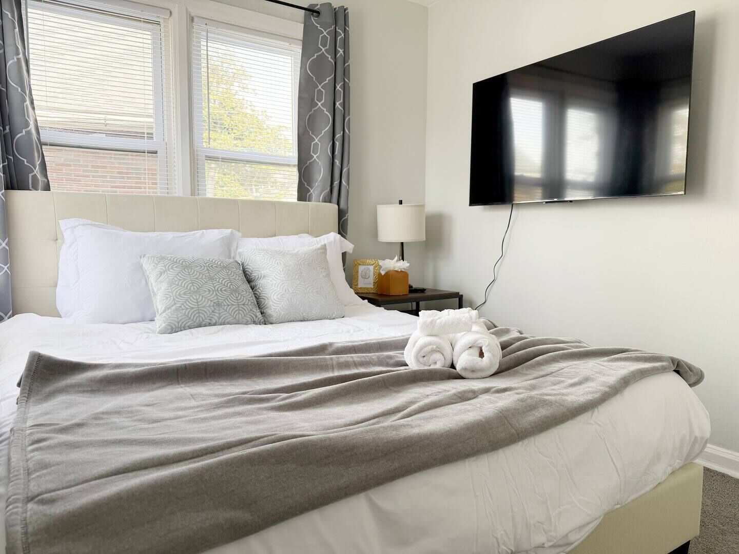 Una cama bien hecha con una manta gris, almohadas blancas y toallas enrolladas. Un televisor de pantalla plana está montado en la pared sobre la cama.