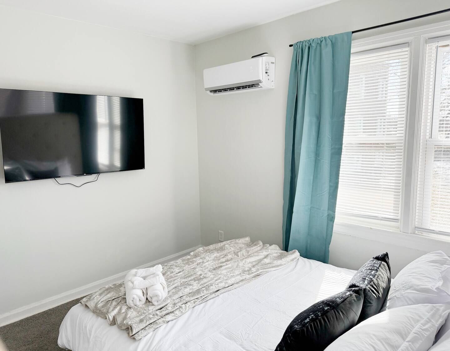 A bedroom with a bed, a flat-screen TV mounted on the wall, an air conditioning unit, and a window with blinds.