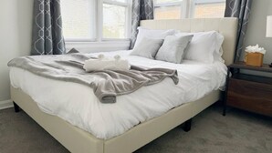 A neatly made bed with white linens and a gray throw blanket, a bedside table with a tissue box, and a window with blinds and patterned curtains. - Riverside A2 - QUEEN 2BD/1BA Apt near River (Evansville)
