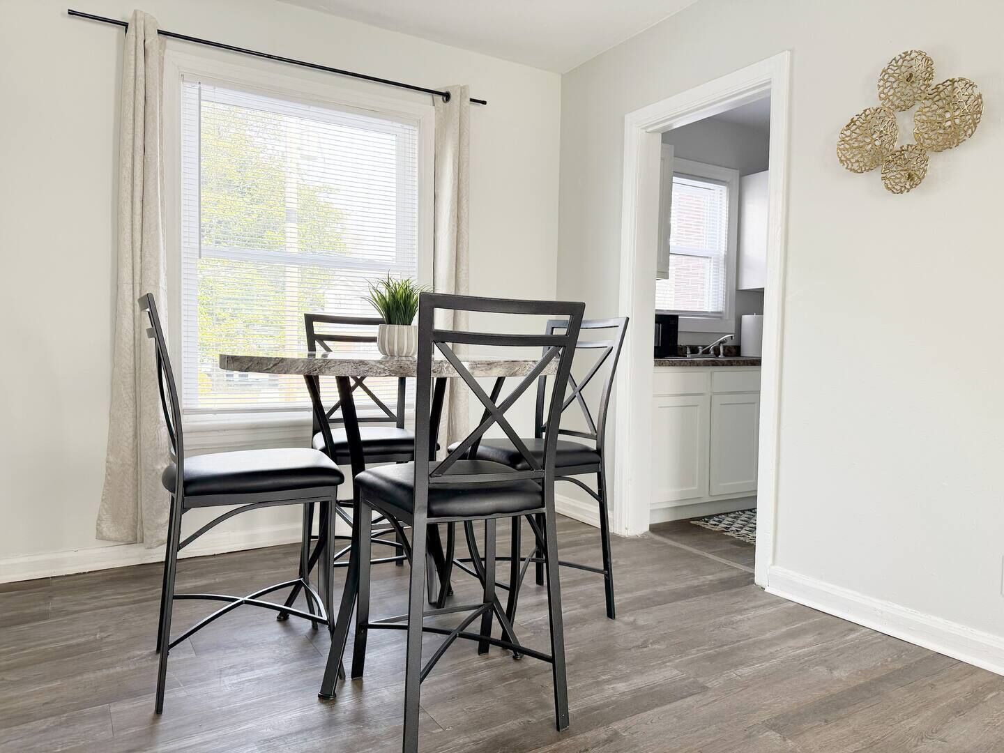 A dining area with a table and four chairs, a window with curtains, and a kitchen visible through an open door.