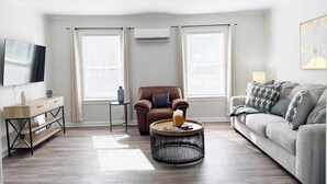 A living room with a brown leather armchair, a grey sofa, a round coffee table, and a TV on the wall. - Riverside A2 - QUEEN 2BD/1BA Apt near River (Evansville)