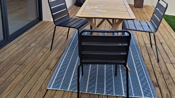 Une terrasse extérieure avec une table en bois et quatre chaises, un barbecue noir et une plante en pot.
