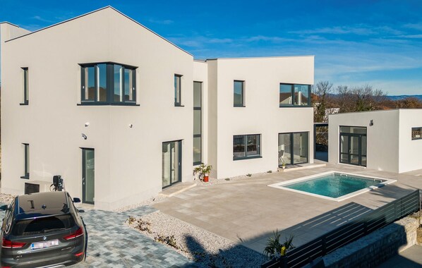 Une maison moderne à deux étages, dotée d’une piscine, avec une voiture garée devant et un ciel dégagé.