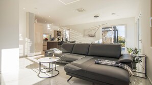 A modern living room with a grey sofa, a small round table, and a glass coffee table. There is a kitchen area in the background with a window and a staircase leading to another level. - Beautiful home in Krk with sauna (Krk)