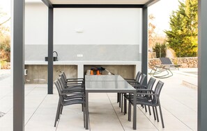 Une terrasse extérieure avec une table rectangulaire et six chaises, un barbecue encastré et un mur de pierres en arrière-plan.