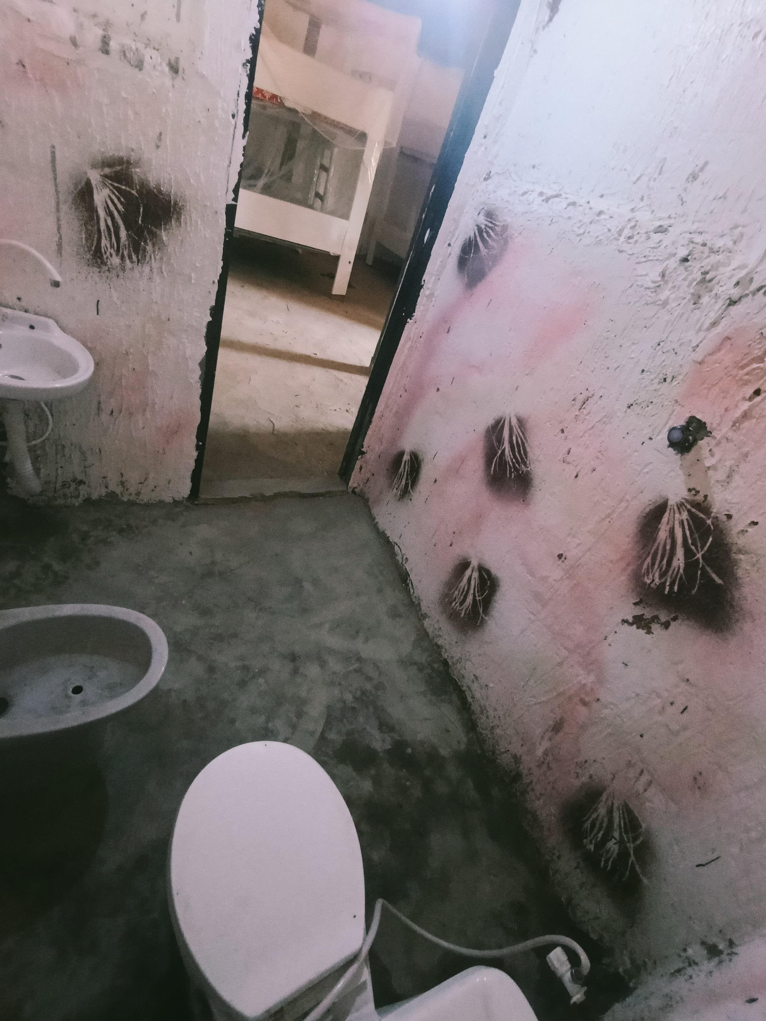 Un baño pequeño y con poca luz, con un inodoro, un lavabo y un espejo. Las paredes tienen una textura y muestran señales de desgaste.