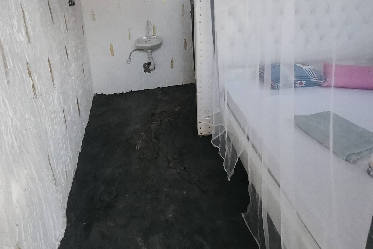 Una habitación pequeña y estrecha con una cama individual cubierta por una sábana blanca translúcida, un lavamanos pequeño y una ventana con cortina estampada.
