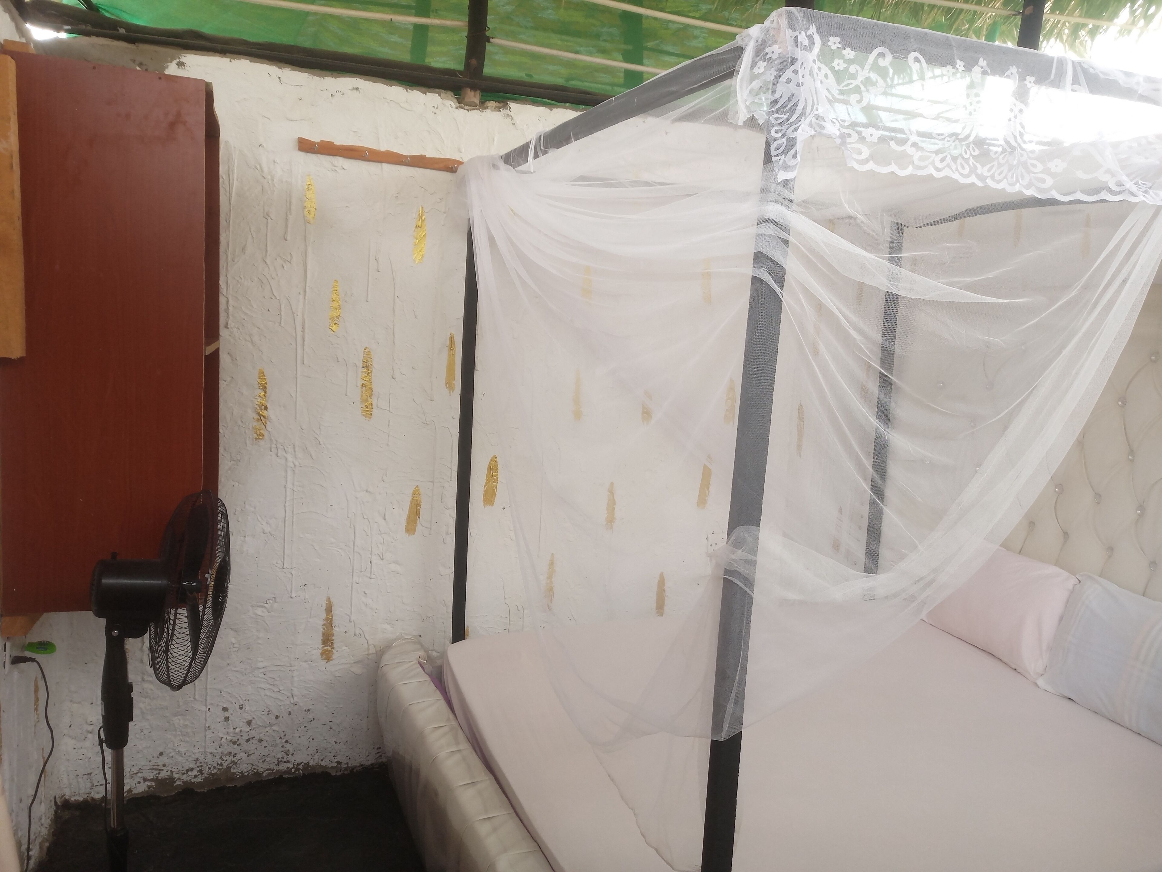 A small, enclosed room with a bed under a mosquito net, a fan, and a wall with gold markings.