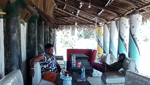 A covered outdoor seating area with cushioned benches and a table, featuring a rustic thatched roof and a view of greenery outside. - Babalao Beach Resort  Backpackers & Tent (Malindi)