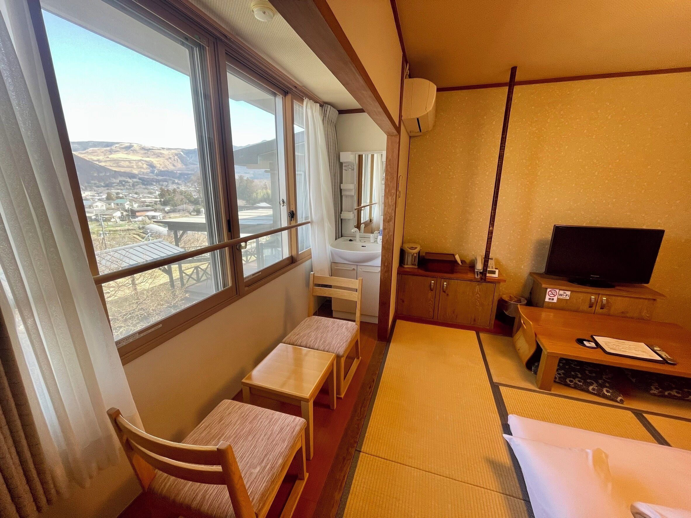 A hotel room with a large window, a small table, a chair, a TV, and a cabinet.