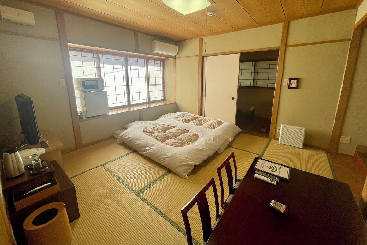A traditional Japanese-style room with tatami flooring, a futon bed, a desk with a computer, and a window with a view of the outside.
