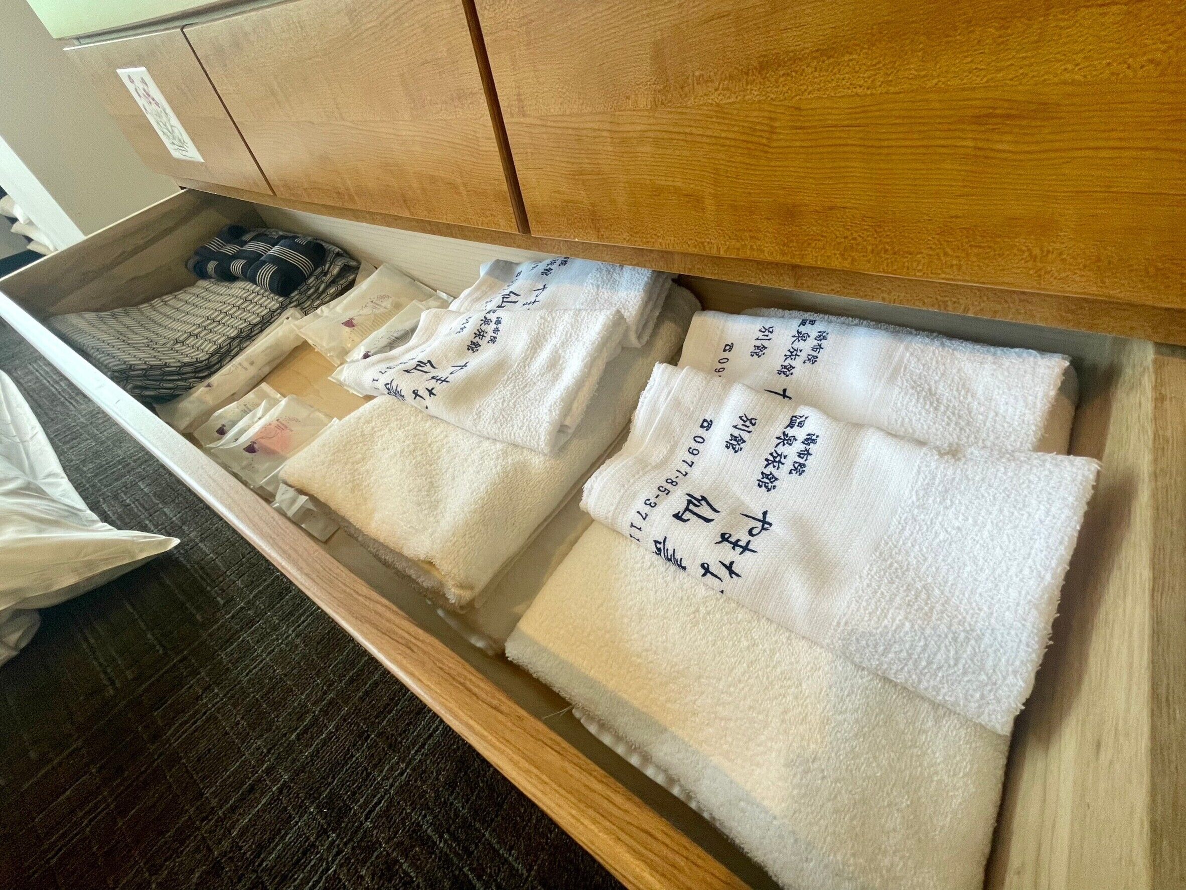 Un cajón con toallas blancas dobladas ordenadamente, algunas con texto impreso, y un par de zapatillas.