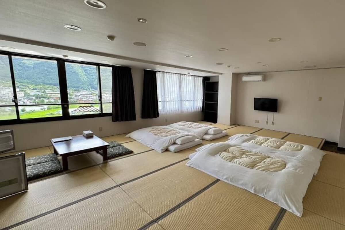 A room with tatami mats, a bed, a desk, a TV, and a window with a view of greenery.