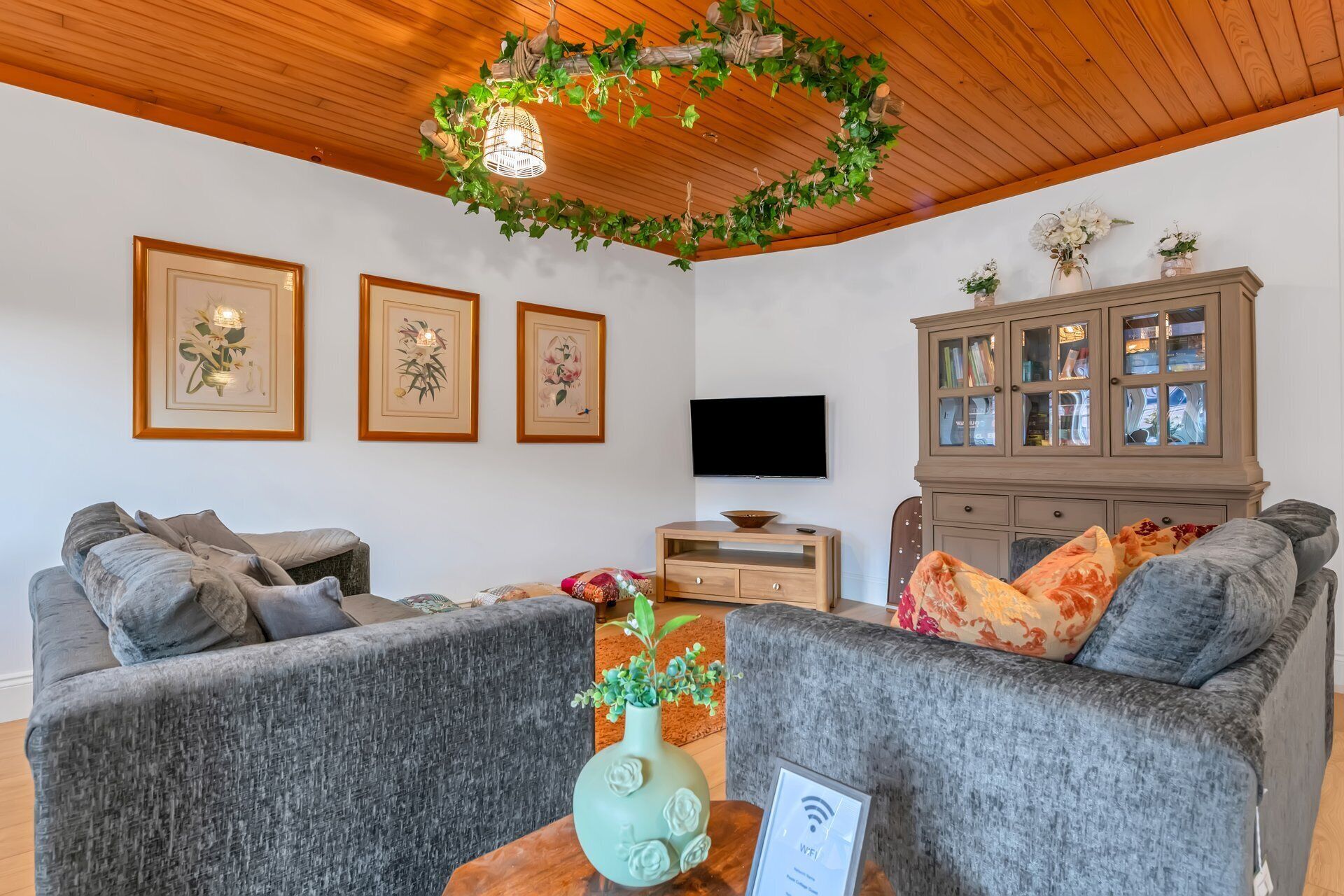 A living room with two grey sofas, a wooden cabinet, a flat-screen TV, and framed floral artwork on the wall.