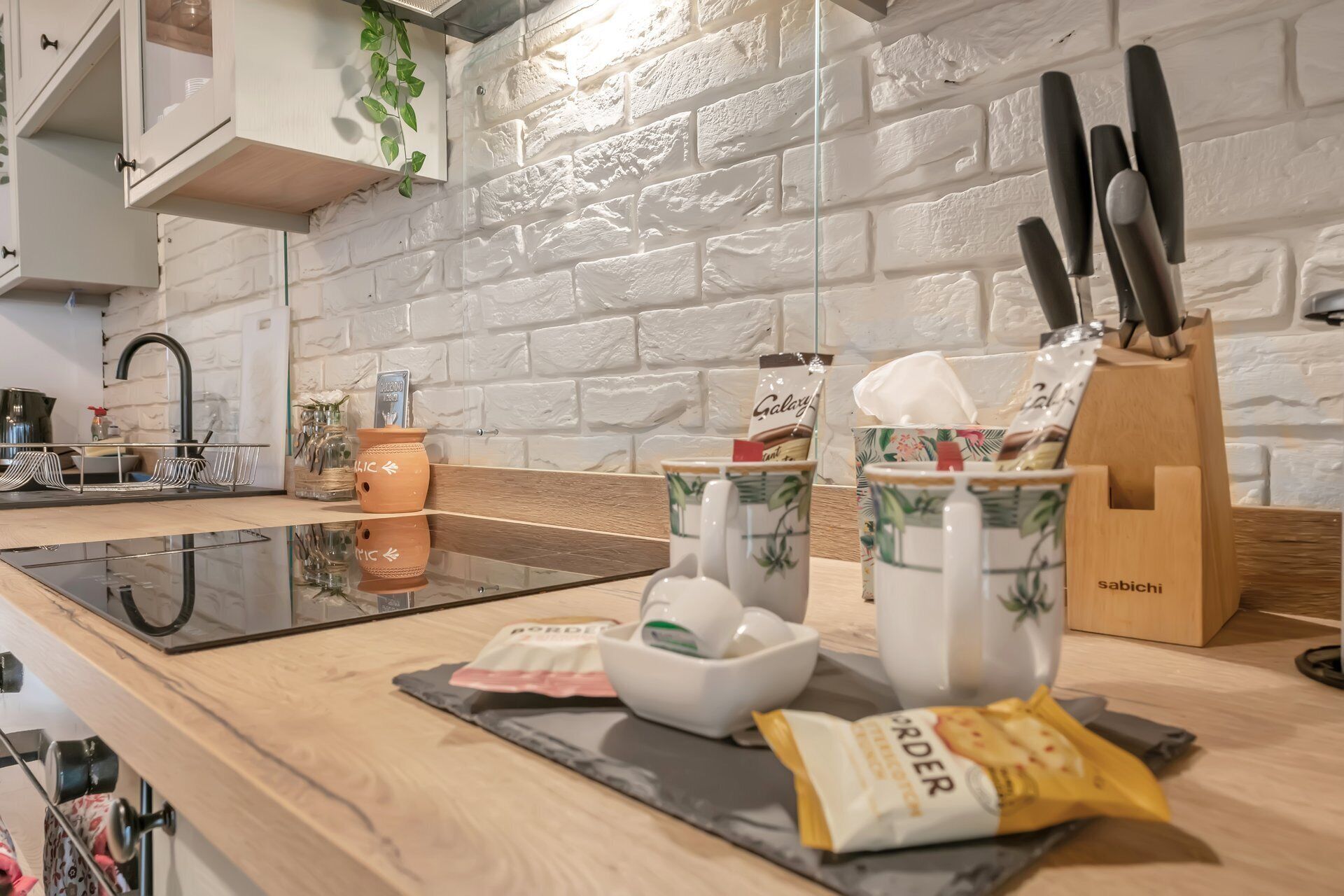 A kitchen with a white brick wall, a wooden countertop, a sink, and various kitchen items including a knife block, a bowl, and a packet of snacks.