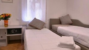 Habitación de hotel con dos camas individuales, un escritorio pequeño con microondas, un jarrón con flores y una ventana con cortinas traslúcidas.