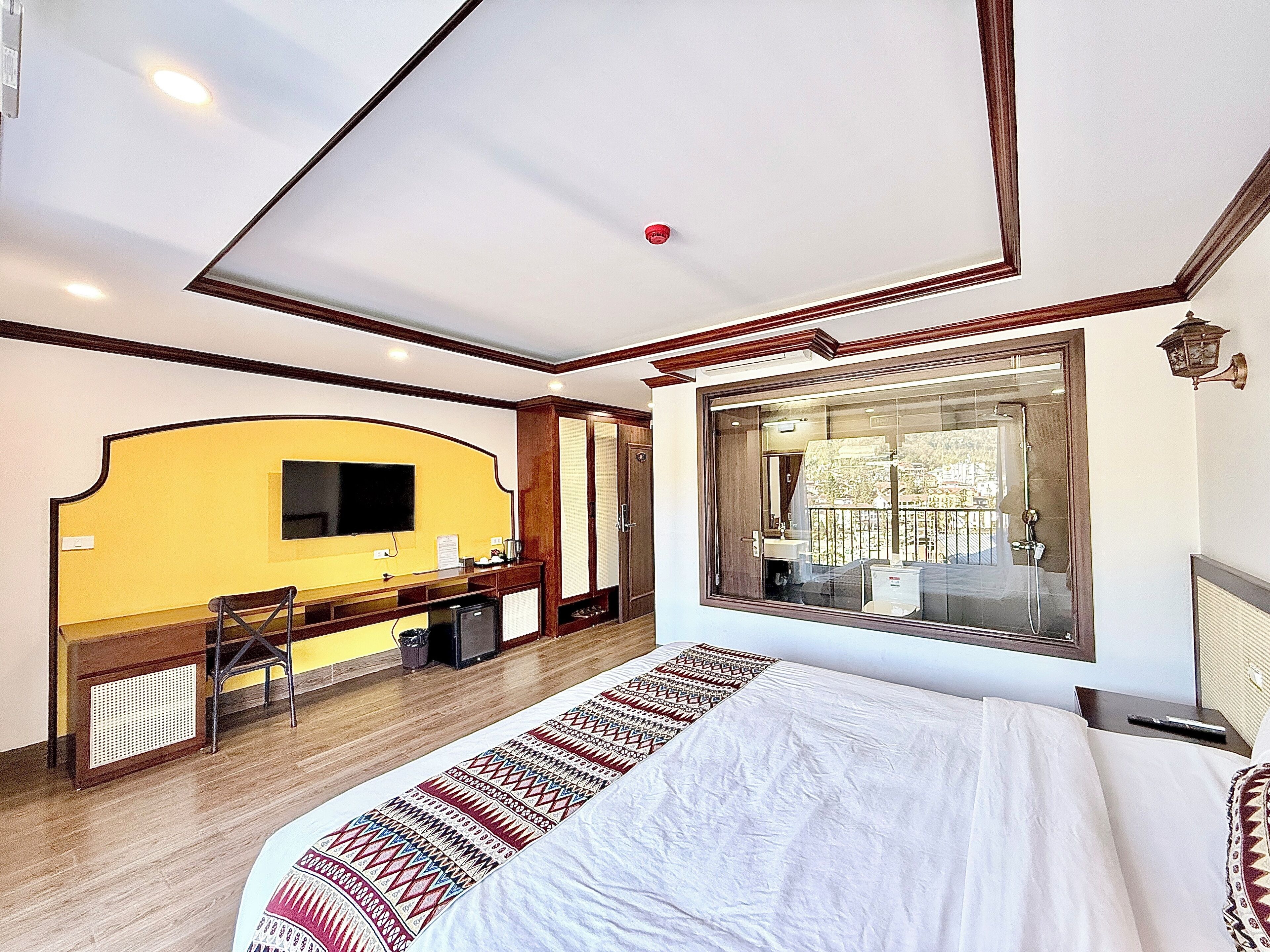 A hotel room with a large bed, a wooden desk, a TV, a bathroom with a window, and a balcony.