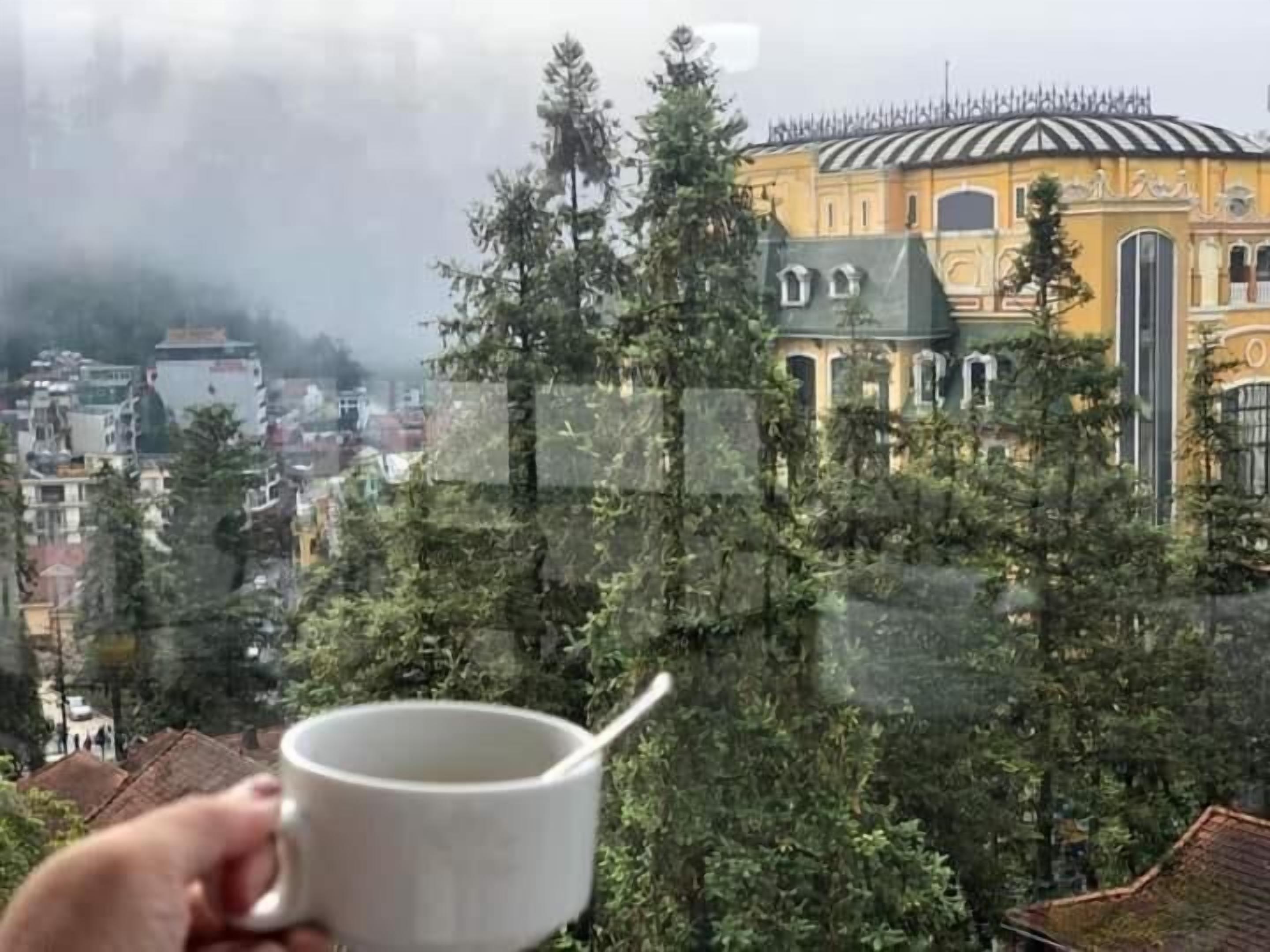 A hand holding a white mug with a spoon, overlooking a cityscape with buildings and trees.