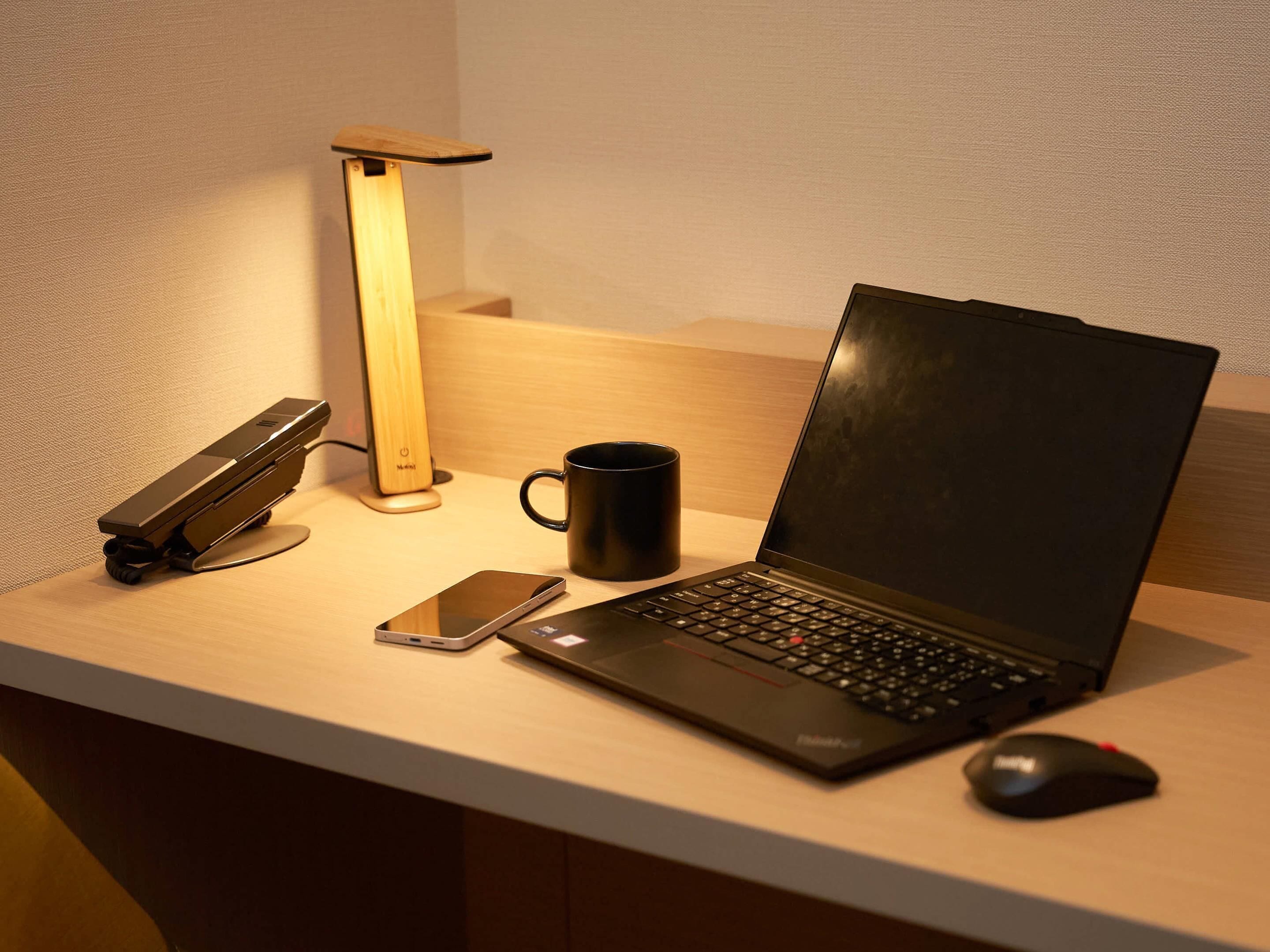 A laptop on a desk with a smartphone, a black mug, a mouse, and a standing lamp.
